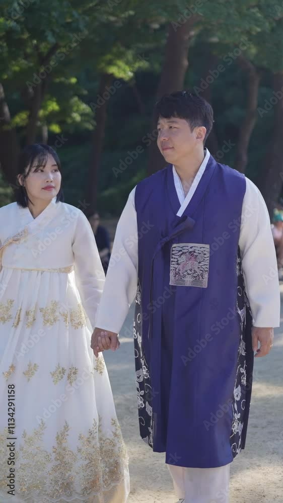 A Korean couple, a man in his 30s and a woman in her 20s, walk together in a park lined with historical buildings, wearing Hanbok from Seoul, South Korea. Slow motion vertical video.