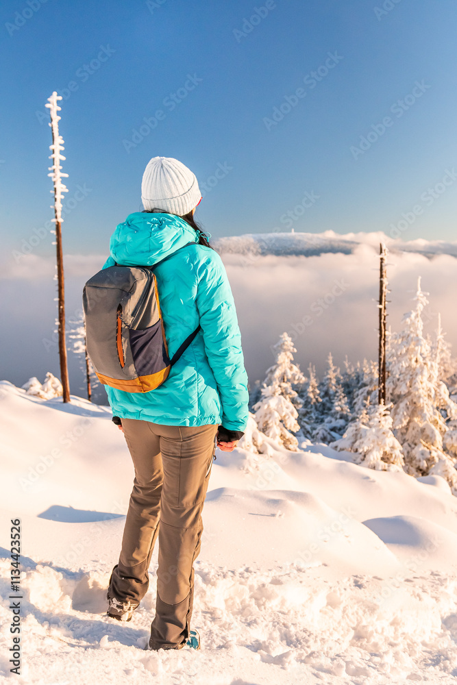 Eastern Sudetes, Young woman hikes on mountain snow-covered trail, walks on white snow over snowy peak and looks at fog in valley, winter landscape in mountains on sunny day.