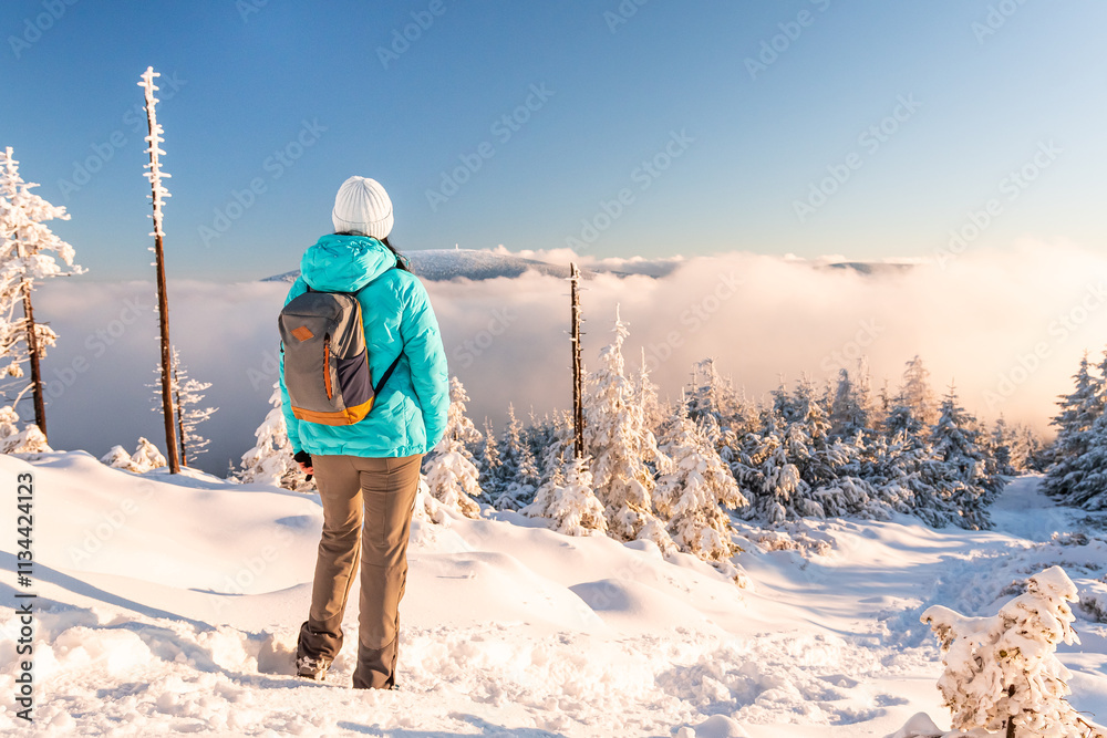 Eastern Sudetes, Young woman hikes on mountain snow-covered trail, walks on white snow over snowy peak and looks at fog in valley, winter landscape in mountains on sunny day.