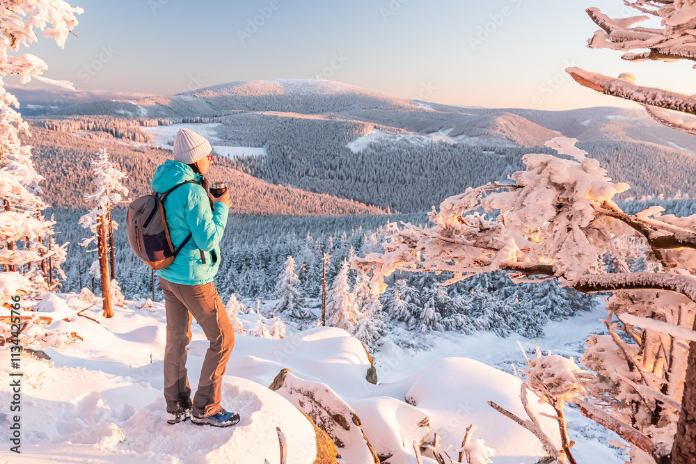 Eastern Sudetes, Young woman hikes on a snow-covered mountain trail, stands on a viewpoint and eats a warm meal, winter landscape in the mountains at sunset.