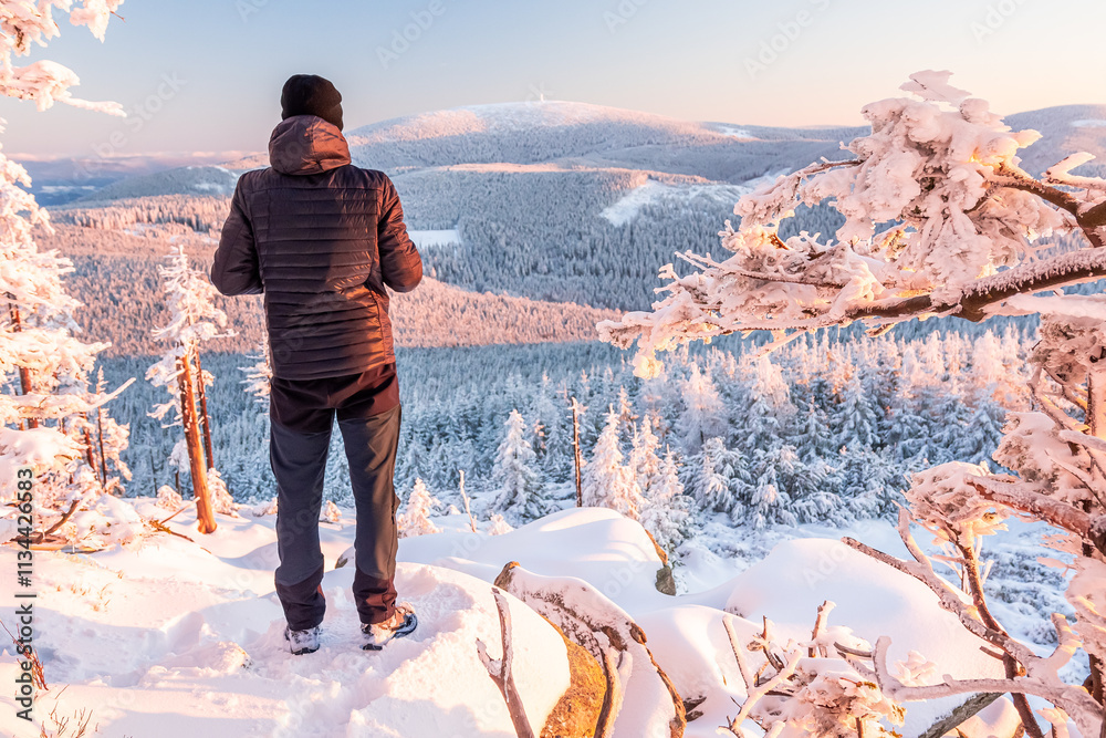 Eastern Sudetes, A young man hikes along a snow-covered mountain trail, stands on a viewpoint and looks at the snow-capped peaks and forest, winter landscape in the mountains.
