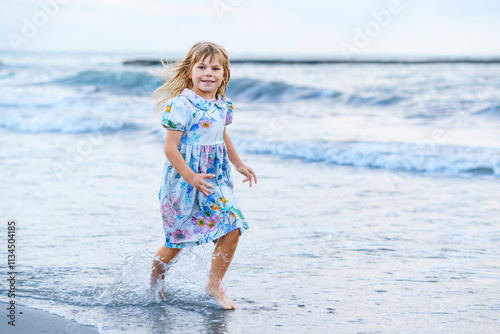 Happy Child, Little Preschool Girl in Dress Running And Jumping In The Waves During Summer Vacation On Exotic Tropical Beach by Sunset. Family Journey On Ocean Coast.