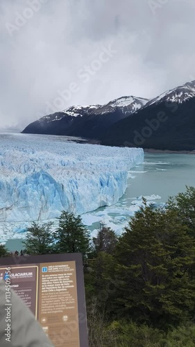 Woman in a grey beanie photographing Perito Moreno Glacier using her smartphone. Shot from a viewing platform with an informational sign in the foreground, blue ice 