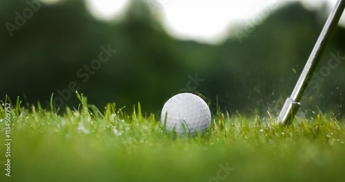 Golf club hits a golf ball in a super slow motion. Drops of morning dew and grass particles rise into the air after the impact.