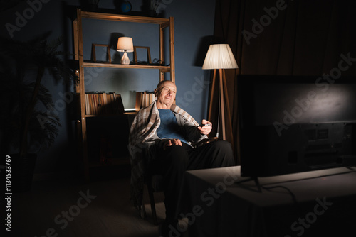 lonely pensioner man sits at home alone in the evening watching TV, calmly with a smile on his face in a chair, holds a remote control in his hands and switches channels.