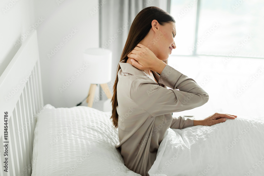 Tired young woman touching aching neck, suffering from pain after uncomfortable sleep, sitting on bed in bedroom at home, side view Tired young woman touching aching neck, suffering from pain after uncomfortable sleep, sitting on bed in bedroom at home, side view