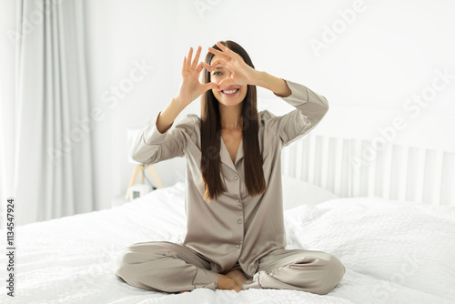 Happy woman wearing comfortable pajamas sitting on white bed and showing heart gesture and smiling at camera photo