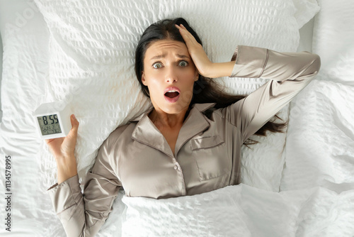 Shocked overslept woman holding alarm clock and touching head, looking at camera with open mouth, lying in bed on her back in morning