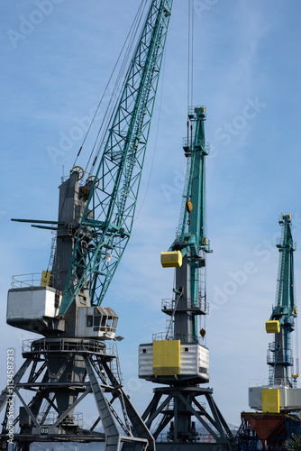 A trio of towering industrial cranes dominates the horizon against a vibrant blue sky