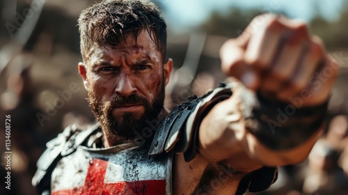 An intense-looking warrior in medieval armor with a red cross appears ready for battle, showcasing strength and determination under a bright blue sky backdrop. photo