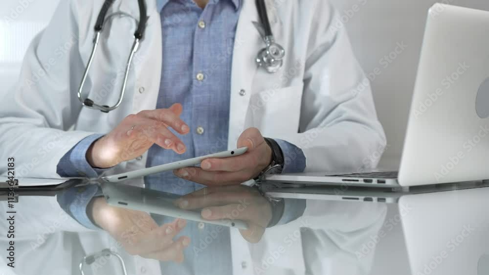 Doctor man, wearing a blue shirt under a medical white coat, is using digital tablet in modern healthcare setting, interacting with patient data, close-up. Medicine concept