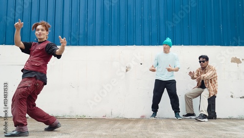 Young street dancer dancing in hip hop style with multicultural friends cheering behind at wall. Attractive handsome man stretch arms at street with blue background. Outdoor sport 2024. Hiphop. photo