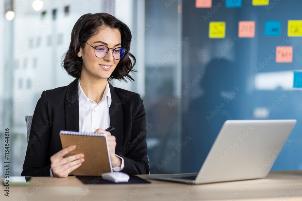 Confident businesswoman with glasses in modern office taking notes. Using laptop and phone for work research. Represents professional women and office work dynamics in business environment.