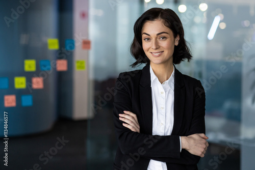 Confident woman in business attire standing with arms crossed in modern office, smiling. Represents leadership, success, and empowerment in professional environment.