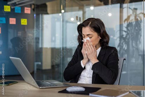 Businesswoman sneezing at office desk with laptop open, showing symptoms of cold. Captures concept of illness at work, stress, and need for better health management during work hours.