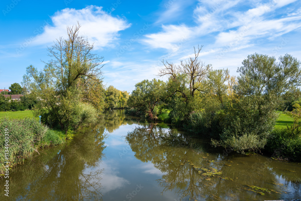 Wornitz river scene in Ebermergen with lush greenery on both banks, reflecting the blue sky and clouds. Trees with varying foliage surround the water Wornitz river scene in Ebermergen with lush greenery on both banks, reflecting the blue sky and clouds. Trees with varying foliage surround the water