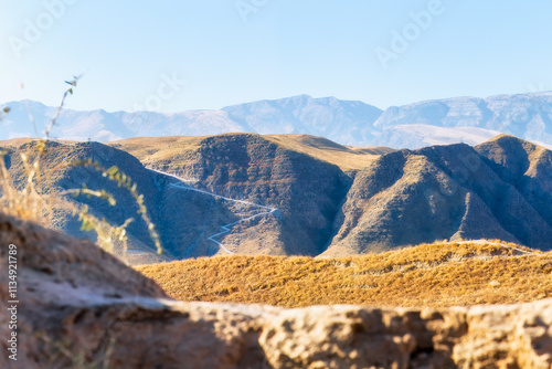 A scenic view of a rugged valley framed by arid terrain and desert vegetation and the majestic Kopet Dag Mountains with higing path in the background under a clear blue sky, Turkmenistan. Copy space