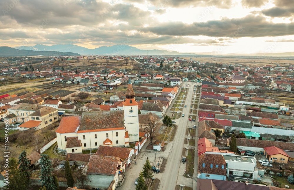 Obraz premium Breathtaking aerial view of Rasnov showcasing quaint rooftops, the historic church, and the stunning backdrop of Piatra Craiului mountains during a serene evening