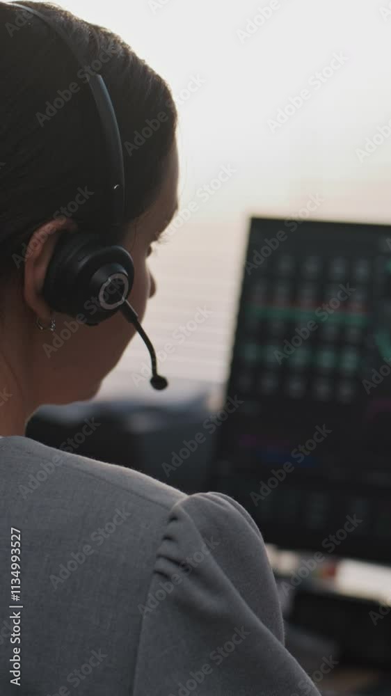 Modern office of International stock exchange company: Back view of female specialist in headset working on computer, communicating with client. PC screen shows equity market and shares in real-time. Modern office of International stock exchange company: Back view of female specialist in headset working on computer, communicating with client. PC screen shows equity market and shares in real-time.