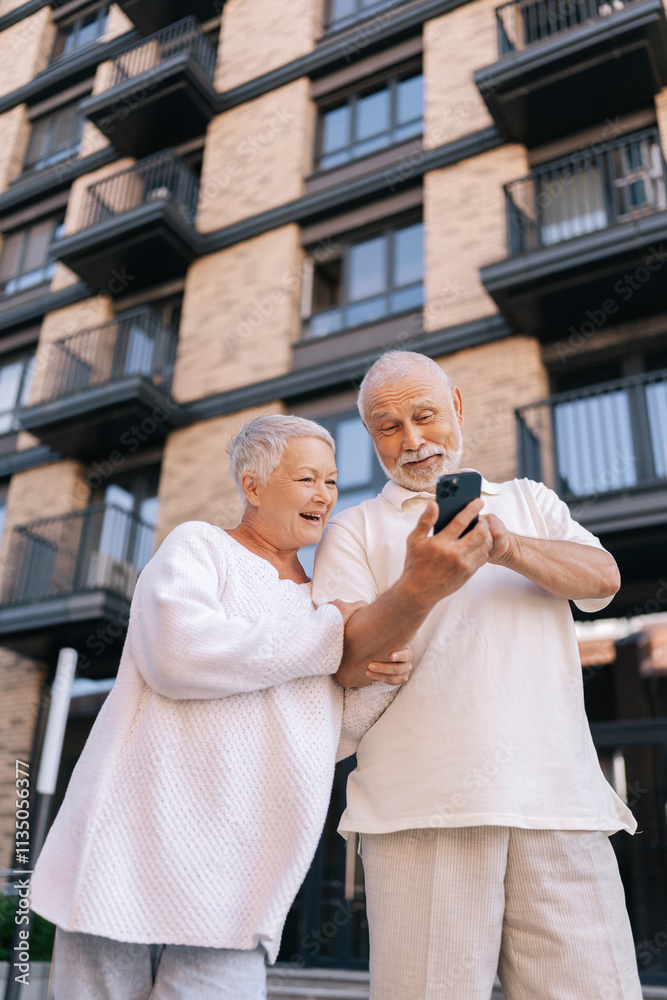 Vertical portrait of cheerful beautiful senior couple with gray-haired in white shirts standing on city street, happily browsing using smartphone, radiating love and joy. Concept of active aging. Vertical portrait of cheerful beautiful senior couple with gray-haired in white shirts standing on city street, happily browsing using smartphone, radiating love and joy. Concept of active aging.