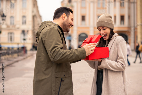 Mid adult Caucasian man surprises his wife with a gift in a European city. The couple is dressed in winter attire, with the man holding a vibrant red present. photo