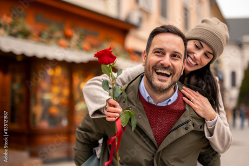 Mid adult Caucasian male carries girlfriend joyfully, holding a rose on International Women's Day. Both dressed in casual winter attire, sharing a moment of happiness on a bustling city street.