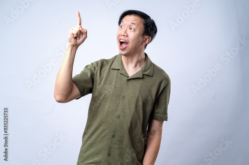 Obraz na plátně An Asian man in a casual green shirt excitedly points upward with one finger, mouth open in expression, standing against a clean white background