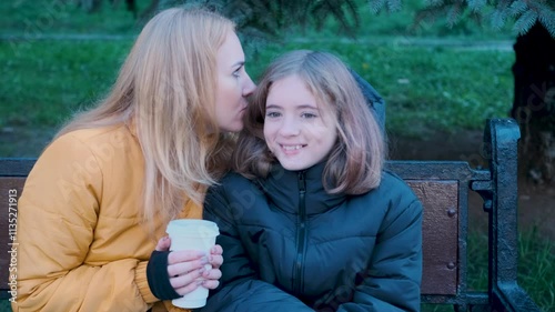 Mother whispers a secret to her teenage daughter while sitting on a bench, capturing a heartfelt moment in slow motion