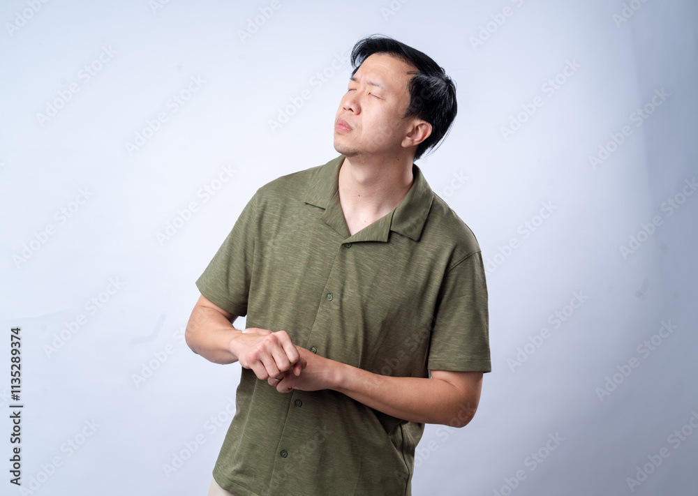 Fototapeta premium An Asian man in a casual green shirt looks to the side with a thoughtful expression, clasping his hands together, standing against a clean white background. The image portrays contemplation