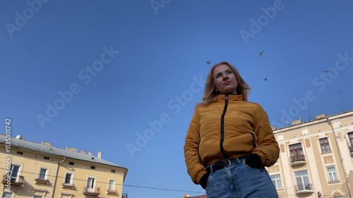Woman in a yellow jacket is standing in a city square, birds flying in the blue sky