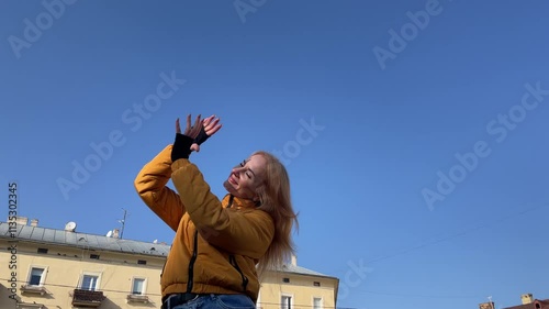 Woman in a yellow jacket raises her hands to the bright blue sky, embracing sunlight with a joyful spirit