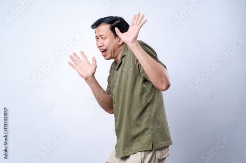 An Asian man wearing a green polo shirt reacts with a startled expression, raising his hands defensively as if in fear or surprise, against a plain white background, showcasing vivid emotion