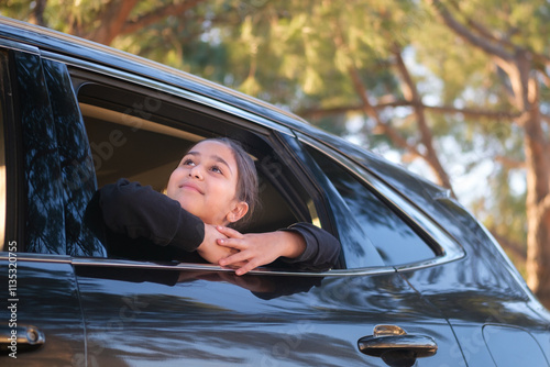 A smiling girl is sitting in a  car by the road . She is looking out from the opened window and enjoying fresh air. 