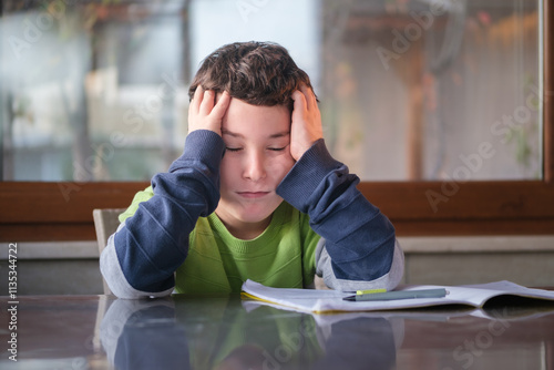 Little boy at home sitting at the table tired doing his homework by himself. Holding head by hand and looking sad.