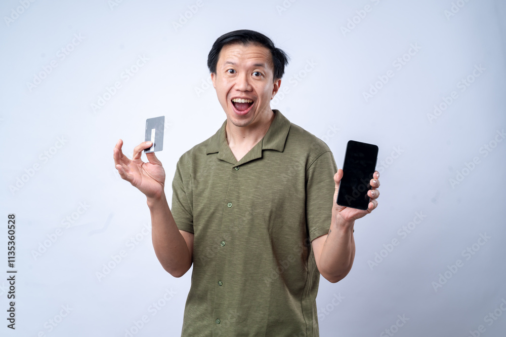 An Asian man in a casual green shirt excitedly looks at his smartphone while holding a credit card, standing against a clean white background. The image portrays online shopping, payment An Asian man in a casual green shirt excitedly looks at his smartphone while holding a credit card, standing against a clean white background. The image portrays online shopping, payment