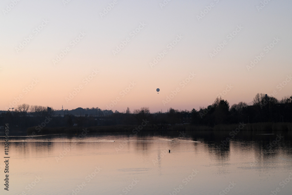 Against the backdrop of a gentle sunset, a hot air balloon is visible in the sky, smoothly rising above the horizon. The water of the lake is illuminated by a soft light Against the backdrop of a gentle sunset, a hot air balloon is visible in the sky, smoothly rising above the horizon. The water of the lake is illuminated by a soft light