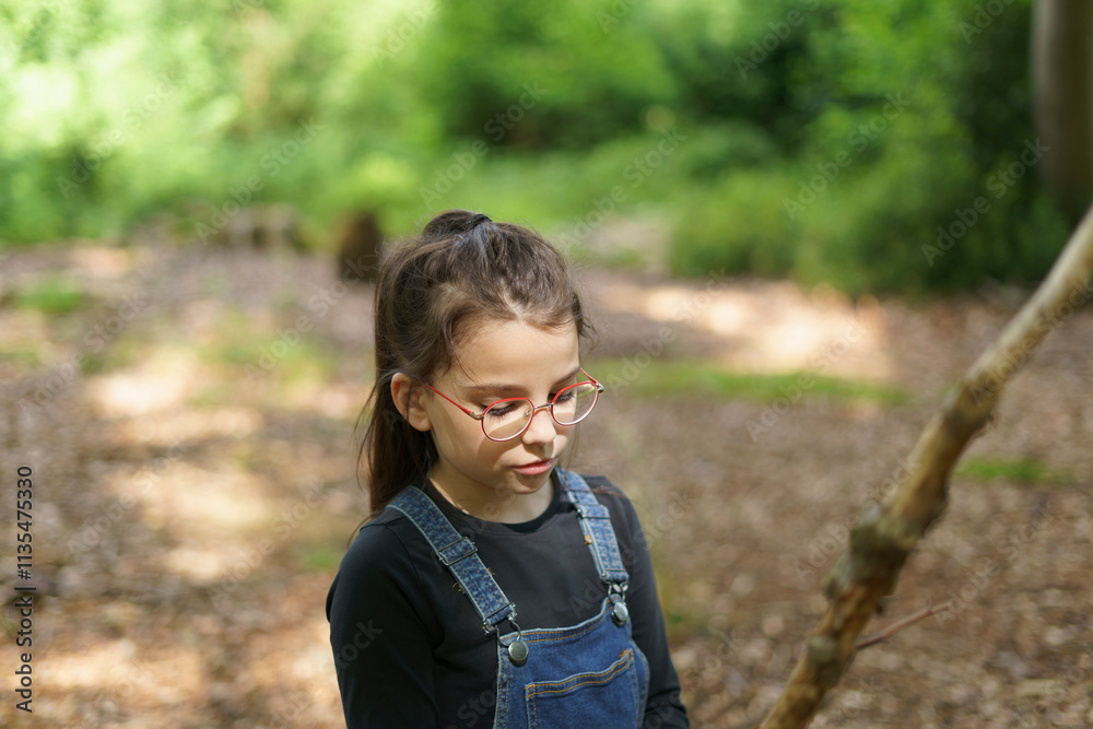 Little cute girl in denim overalls and glasses builds a house of sticks in the forest in summer. Concept of children's fun, outdoor games Little cute girl in denim overalls and glasses builds a house of sticks in the forest in summer. Concept of children's fun, outdoor games