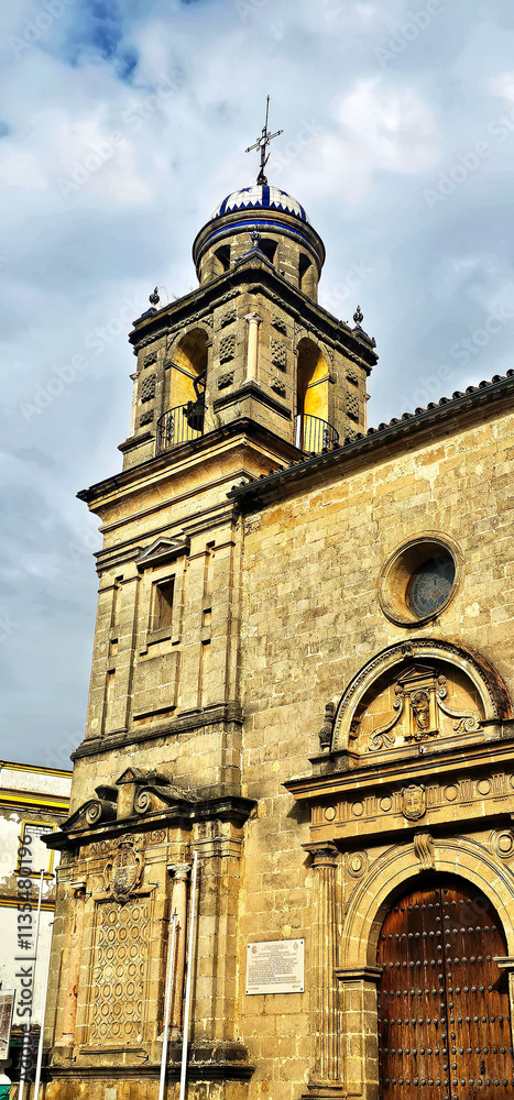 Fototapeta premium Church facade with a stone bell tower topped by a blue and white dome; detailed carvings on the arched wooden door and window frame; cloudy sky above;
