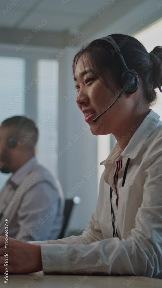 Call center office: Vertical shot of tired, stressed Asian hotline operator in headset using computer, finishing phone call with client, holding head. Team of diverse customer assistance service staff Call center office: Vertical shot of tired, stressed Asian hotline operator in headset using computer, finishing phone call with client, holding head. Team of diverse customer assistance service staff
