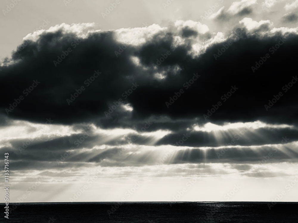 Dramatic black and white photo of sunbeams piercing through dark, stormy clouds over a calm ocean. Dramatic black and white photo of sunbeams piercing through dark, stormy clouds over a calm ocean.