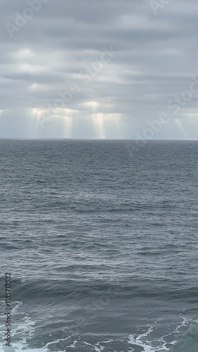 Sunlight beams cut through a cloudy sky, illuminating the calm ocean surface below.