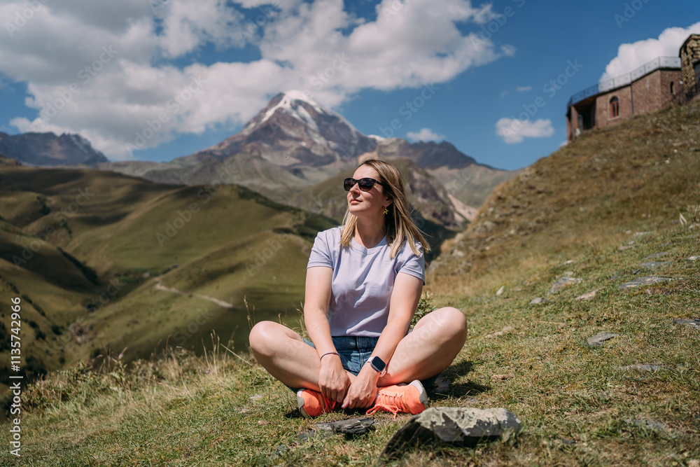 A girl is sitting in a lotus position against the background of Mount Kazbek in Georgia. The girl is wearing a T-shirt, denim shorts, sunglasses. A mountain in a snow cap