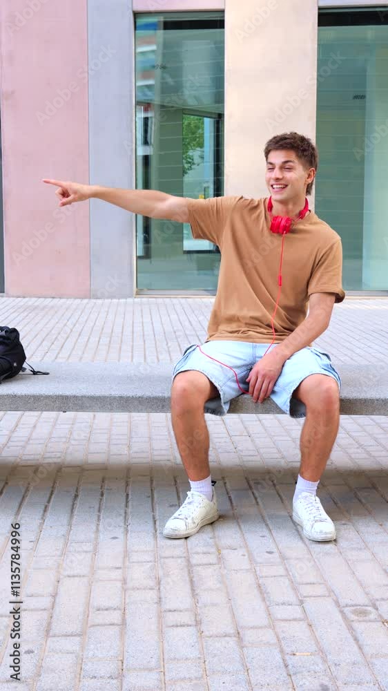 A young man sits on a concrete bench outdoors, wearing red headphones and casual attire. He gestures by pointing to the side, appearing engaged and expressive in a modern urban setting.Vertical A young man sits on a concrete bench outdoors, wearing red headphones and casual attire. He gestures by pointing to the side, appearing engaged and expressive in a modern urban setting.Vertical