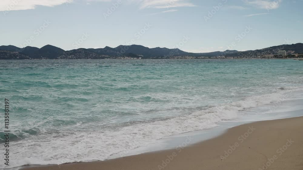Slow motion Clear blue waves with strong wind on a sandy empty beach in Cannes with Theoule-sur-Mer on background Slow motion Clear blue waves with strong wind on a sandy empty beach in Cannes with Theoule-sur-Mer on background