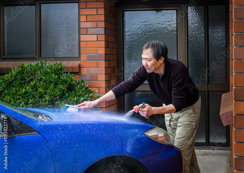 Obraz na płótnie Man washing a blue car in front of the house using a brush and water spray from a hose. w salonie
