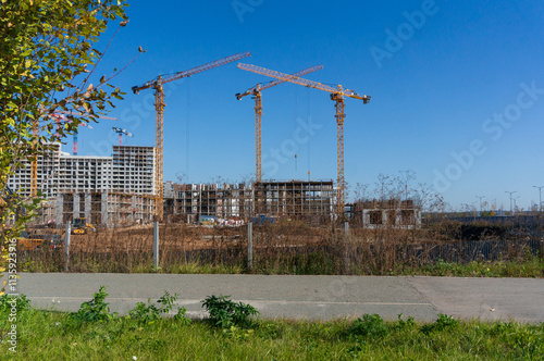 Construction of a multi-story building with cranes against a blue sky, development of the outskirts of the city, expansion of the city