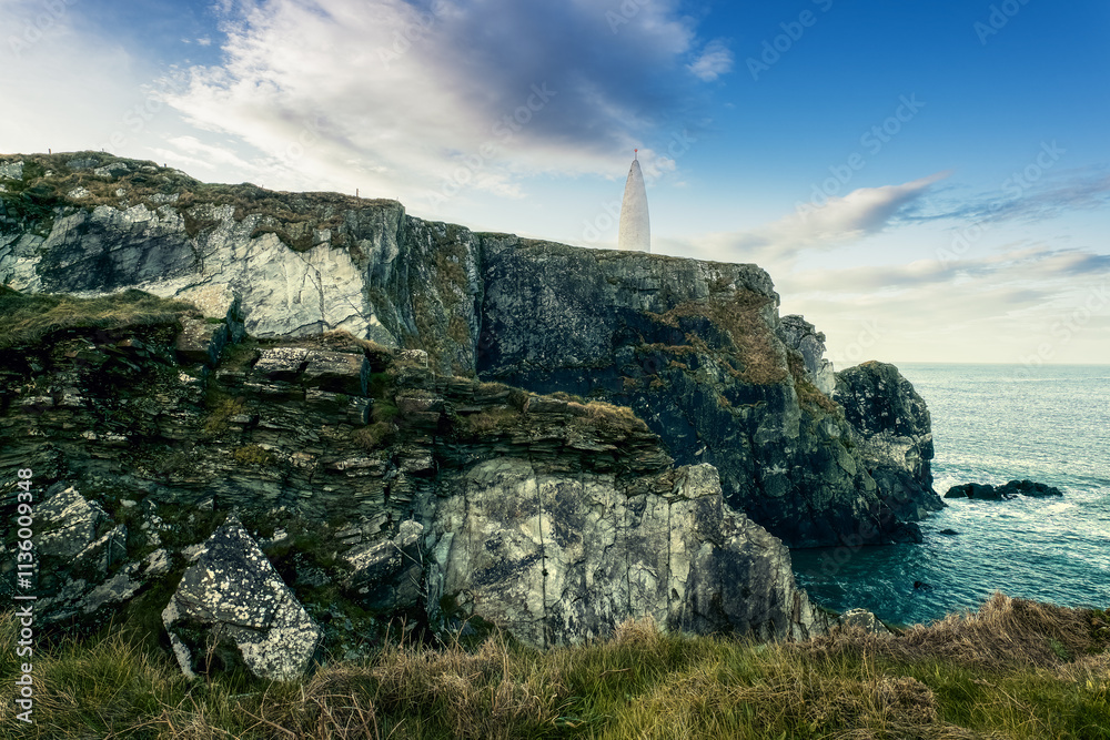 Fototapeta premium A lighthouse stands tall on a rocky cliff overlooking the ocean, surrounded by grassy terrain. The sky is partly cloudy, adding a serene atmosphere to the coastal setting.