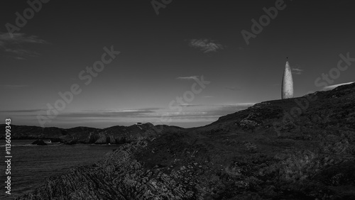 A white, tall, slender marker stands on a grassy cliff. Dark, rugged rocks form the cliff face. Sea stretches to a distant coastline. photo