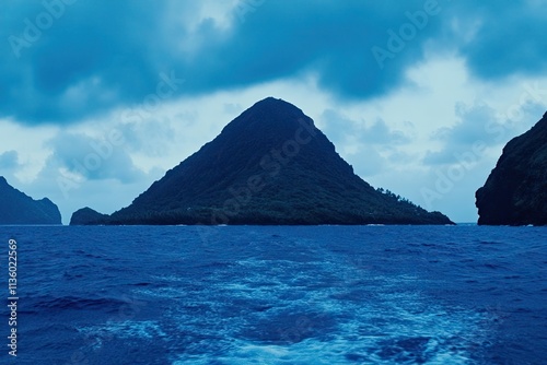 Dramatic ocean landscape featuring a majestic, dark, mountainous island under a brooding sky. The deep blue water creates a tranquil yet powerful mood.