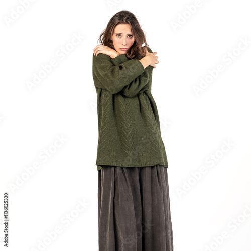 A woman in a green oversized sweater and gray skirt poses against a white background. Model hugging herself with arms